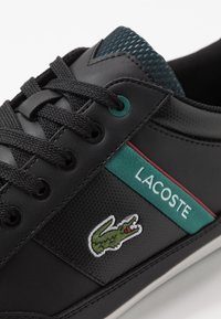 Musta lenkkikenkä, jossa verkko- ja synteettisiä materiaaleja, vihreä ja punainen korostevyö, jossa lukee "LACOSTE", sekä vihreä krokotiililogo.