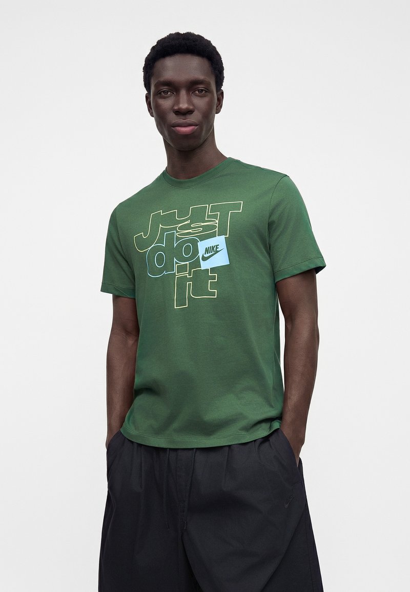 Ung mand iført en grøn Nike t-shirt med teksten "Just do it" og et blåt Nike logo-mærke, kombineret med sorte shorts.