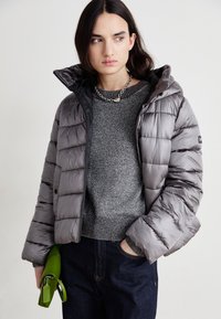 Doudoune grise avec capuche, présentant un design matelassé et des accents noirs. Portée par-dessus un pull gris scintillant et associée à un jean foncé et un sac à main vert.