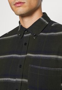 Donkergroene knoopshirt met kraag, voorzien van zwarte knopen en witte strepen. Inclusief een zak en gestructureerde stof.