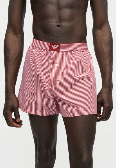 Αρσενικός κορμός που φορά κόκκινα και άσπρα κάθετα ριγέ μποξεράκια με ελαστική μέση με επώνυμο λογότυπο και κουμπωτό κλείσιμο, στέκεται μπροστά σε απλό φόντο.