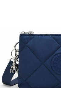 Borsa a tracolla trapuntata blu navy con logo rotondo in rilievo, zip argentata e un piccolo portachiavi a forma di scimmia argentata attaccato alla tracolla.