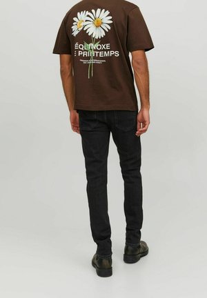 Mężczyzna ubrany w brązowy t-shirt z kwiatami stokrotki i francuskim napisem na plecach, w czarnych jeansach i ciemnobrązowych butach, stojący na szarej podłodze.