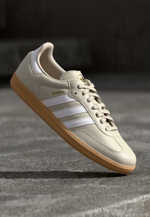 Beige Adidas Samba sneaker med hvide striber og gummisål, vinklet over en tekstureret grå overflade.