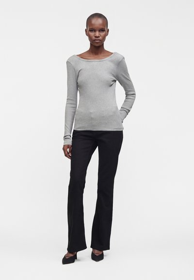 Femme debout sur fond blanc portant un haut à manches longues gris clair, un pantalon noir et des chaussures noires à talons.