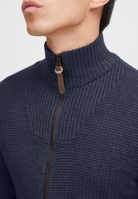 Gilet zippé bleu marine foncé avec col haut côte, corps en tricot texturé, fermeture éclair marron contrastante et design angulaire sur la poitrine.
