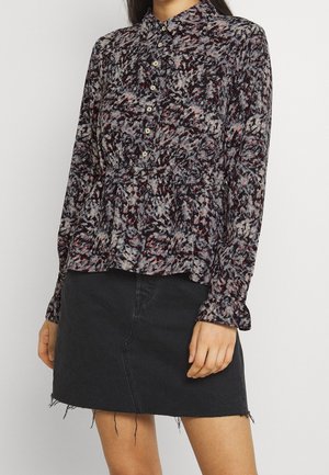 Femme portant un chemisier à manches longues boutonné avec un imprimé abstrait noir, rouge et blanc, rentré dans une jupe en denim noire effilochée.