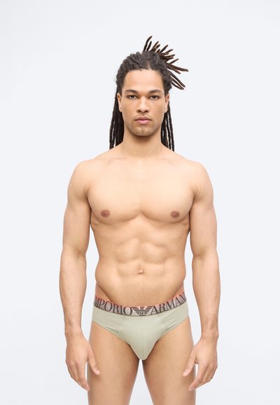 Μυώδης άνδρας με κοτσίδες ντρέντλοκς, φορώντας ανοιχτό πράσινο εσώρουχο Emporio Armani, στέκεται μπροστά σε λευκό, απλό φόντο.
