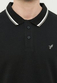 Άνδρας που φοράει μαύρο πόλο πουκάμισο με δύο κουμπιά, λευκό και γκρι ριγέ γιακά και μικρό γκρι λογότυπο πουλιού στο στήθος.