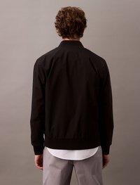 Black bomber jacket with a ribbed collar, cuffs, and hem. Smooth texture with a zip closure, fitted design, suitable for layering over shirts.