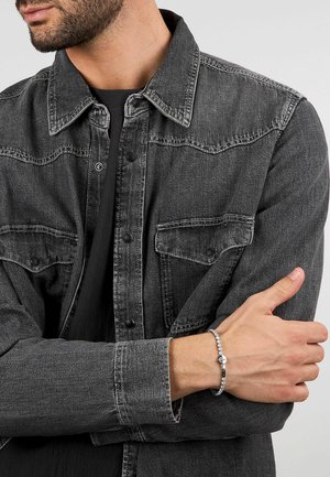 Uomo che indossa una giacca di denim nera e una maglietta nera, con le braccia incrociate, che mostra un braccialetto a catena d'argento al polso sinistro.