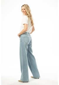 Helle blaue weite Jeans aus Denim mit hoher Taille, hinteren Taschen und einem rohen Saum. Kombiniert mit einem weißen T-Shirt und beigen Schuhen.