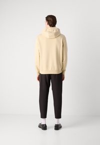 Sudadera beige con capucha ajustable, corte relajado, puños acanalados, combinada con pantalones negros tipo tapered y zapatos negros. Textura suave en general.