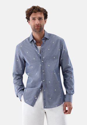 Mann trägt ein blaues Hemd mit Knöpfen und kleinen weißen Palmenmotiven sowie weiße Hosen, steht vor einem schlichten hellen Hintergrund.