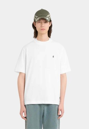 Jeune personne portant un T-shirt blanc oversize avec un petit logo É noir et une casquette verte ornée de motifs de crânes blancs.