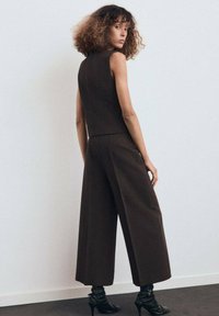 Haut sans manches marron associé à un pantalon large, tous deux en tissu texturé. Des bottes à talons noires complètent la tenue.