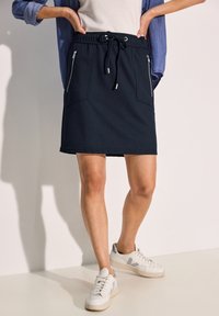 Jupe de sport bleu marine avec une taille à cordon, dotée de deux poches zippées. Fabriquée dans un tissu lisse et extensible avec une longueur allant jusqu'au genou.