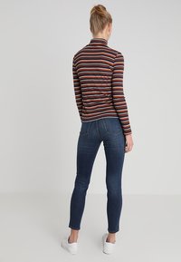Camiseta de manga larga a rayas en rojo oscuro, naranja y crema, combinada con jeans ajustados de color azul oscuro y zapatillas blancas. Vista por la parte de atrás.