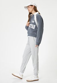Pullover grigio con colletto bianco a zip e logo, abbinato a pantaloni jogger grigi chiari e sneaker bianche con suola marrone; ensemble di abbigliamento attivo casual.