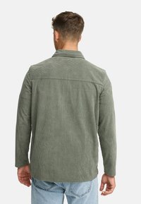 Mand med kort brunt hår iført en grøn kordfløjlsk jacket og lyseblå jeans, vist fra ryggen mod en ensfarvet baggrund.