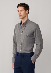 Hombre vestido con una camisa gris de botones con un pequeño logo bordado en el pecho y pantalones azul marino, de pie con una mano en el bolsillo.