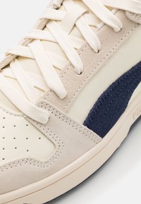 Gros plan sur une basket crème avec des superpositions en daim beige, des lacets blancs et une bande latérale en daim bleu marine sur une semelle blanche.