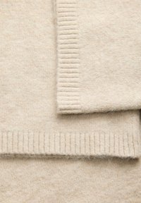 Manica di un morbido maglione di lana beige che si sovrappone al corpo, con polsini e orlo a coste, mostrando texture e dettagli del punto.