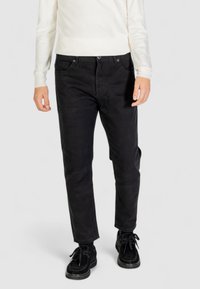 Pantaloni neri in cotone a gamba slim, con tasche frontali e chiusura con bottone, abbinati a una camicia a maniche lunghe color crema e scarpe nere.