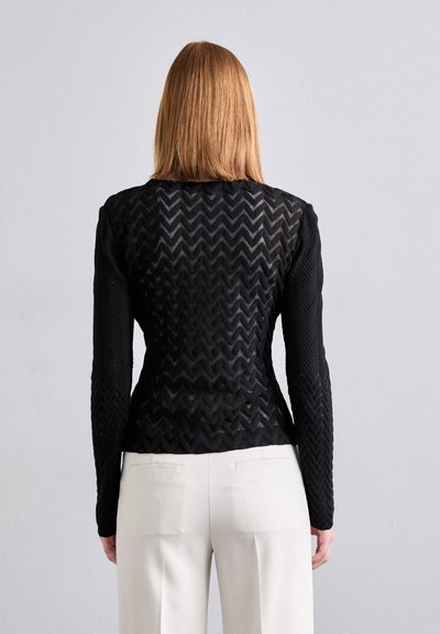 Haut noir à manches longues avec un motif zigzag texturé, dos transparent et coupe ajustée. Associé à un pantalon de couleur claire.