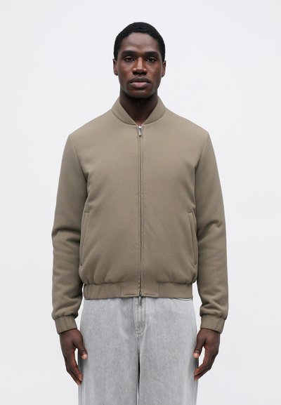 Veste bomber kaki avec une finition texturée, fermeture éclair à l'avant, poignets et ourlet côtelés. Associée à un pantalon gris clair à coupe décontractée.