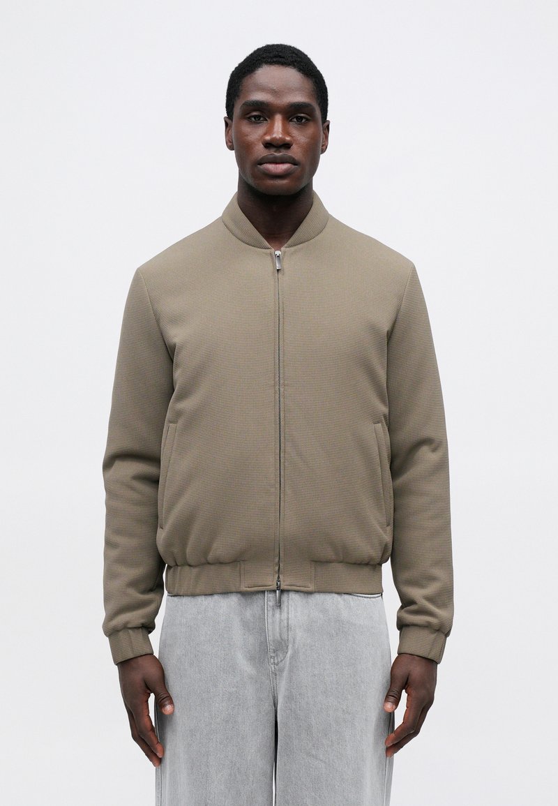 Khaki bomber jacket με υφή, μπροστινό φερμουάρ, ριμπ μανσέτες και στρίφωμα. Συνδυασμένο με ανοιχτό γκρι παντελόνι σε άνετη γραμμή.