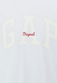 White cotton t-shirt featuring large white letters "GAP" and the word "Original" in red cursive, with a smooth texture and casual design.