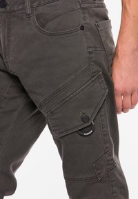 Cargo-broeken van donkergrijze stof kenmerken zich door meerdere zakken, een ritssluiting met klep en een ronde hardware-accent met het label "FASHION."