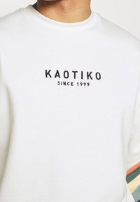Felpa bianca con il logo "KAOTIKO" e "SINCE 1999" in ricamo nero, con colletto a costine e tessuto testurizzato.