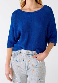 Niebieski sweter z dzianiny o szerokim dekolcie i krótkich rękawach, w połączeniu z jasnymi dżinsami w splatterowe wzory w kolorze pomarańczowym i białym.