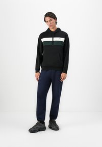 Ung mann iført svart hettegenser med hvite og grønne striper, marineblå treningsbukser og sorte sneakers, stående mot en hvit bakgrunn.