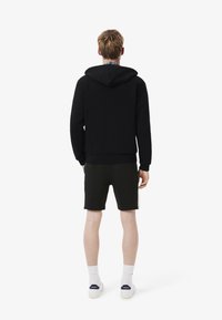 Zwarte hoodie met een trekkoord, gedragen over een geruit shirt, gecombineerd met zwarte shorts en witte sokken, met een minimalistisch ontwerp.