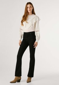 Blusa blanca con cuello y mangas con volantes, combinada con vaqueros negros de campana y botines de tacón en color beige. Tela suave y diseño ajustado.
