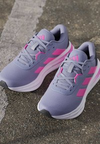 Baskets de sport grises avec des accents roses, tige en maille texturée, épaisse semelle intermédiaire blanche et embout en caoutchouc noir. Logo avec trois bandes visible.