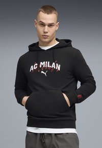 Sweat à capuche noir avec logo "AC MILAN" blanc et accents rouges, doté d'une poche avant et de poignets côtelés, porté sur une chemise blanche.