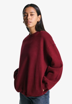 Egy hosszú, sötét hajú nő bordó, oversized pulóvert visel, kezét a zsebében tartja, egy sima fehér háttér előtt.