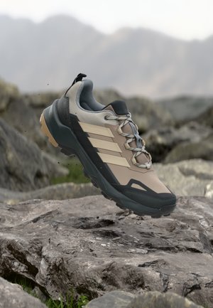 Beiger und grauer Wanderschuh mit schwarzer Sohle und Streifen, schwebend über felsigem Gelände mit verschwommenem Berg-Hintergrund.