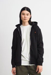 Schwarze, mit einem Reißverschluss versehene Kapuzenjacke aus Baumwolle mit einer Vordertasche und Kordelzugkapuze, getragen über einem hellen, gesprenkelten Shirt. Strukturierte Stoffdetails sind sichtbar.