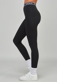 Modellen poserar sidledes iförd svarta leggings och vita sneakers med synliga strumpor. Midjebandet har varumärket SIKSILK.