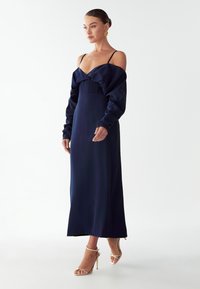 Dlouhá saténová navy modrá šaty s ramínky bez rukávů, nařaseným korzetem a rovnou sukní. Modelka má na sobě sandály na vysokém podpatku v tělové barvě.
