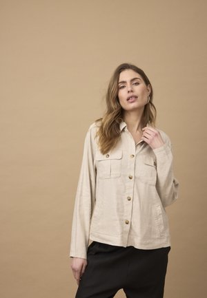 Femme aux cheveux bruns ondulés portant une chemise beige à boutons avec poches et un pantalon noir, debout devant un fond beige uni.