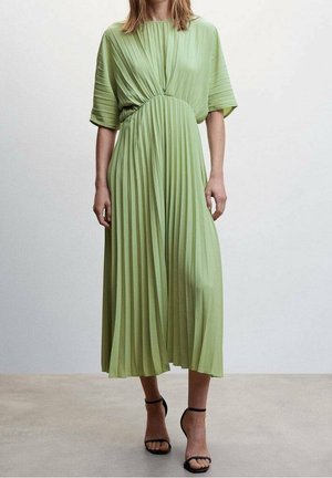 Kobieta ubrana w jasnozieloną plisowaną sukienkę midi z szerokimi krótkimi rękawami oraz czarne sandały na wysokim obcasie z paskiem wokół kostki, stojąca na neutralnej podłodze.
