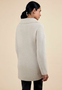 Beige kötött pulóver széles, hajtott gallérral, hosszú ujjakkal és bordázott mintázattal; a csípő alatt végződik, és oldalsó hasítékokkal rendelkezik a kényelmes viselet érdekében.