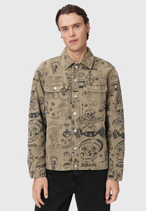 Jonge man draagt een beige jas met knopen met zwarte tatoeage-achtige schetsen en zinnen, gecombineerd met een zwarte broek, tegen een neutrale achtergrond.