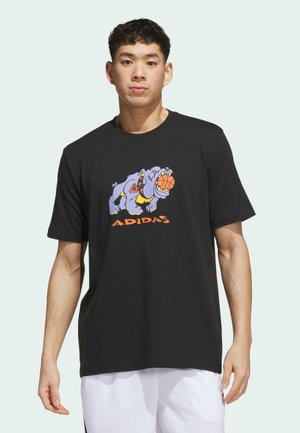 Giovane uomo che indossa una t-shirt nera Adidas con una grafica di un cane viola che morde un pallone da basket.